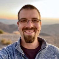 Man with glasses smiles in front of mountains and a sunset